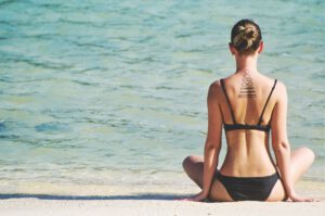 Vrouw in bikini op het strand in kleermakerszit, met de rug naar de camera